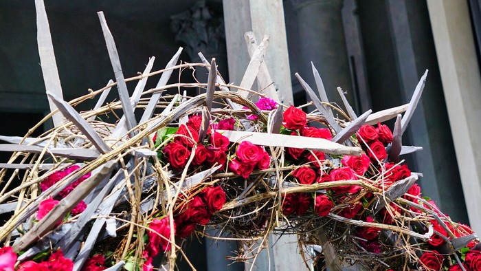 Rosenkranz aus roten Rosen, Stroh und übergroßen, aus Holz geschnitzten Dornen. Über dem Rosenkranz steht ein Kruzifix. 