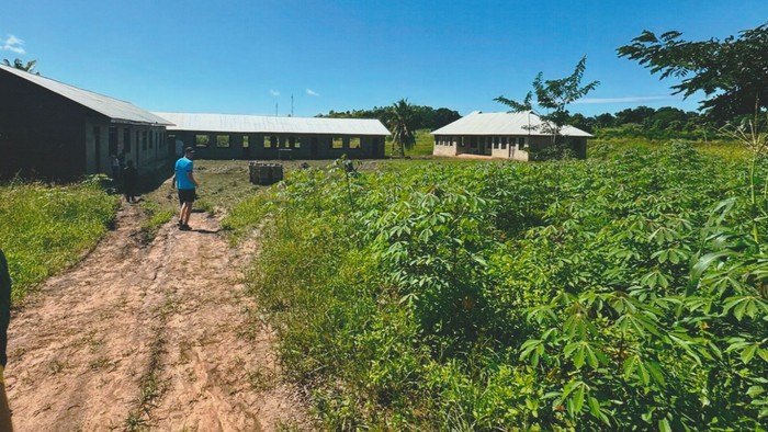 Eine Person geht durch ein üppiges grünes Feld nahe einfachen, langen Gebäuden unter einem klaren blauen Himmel.