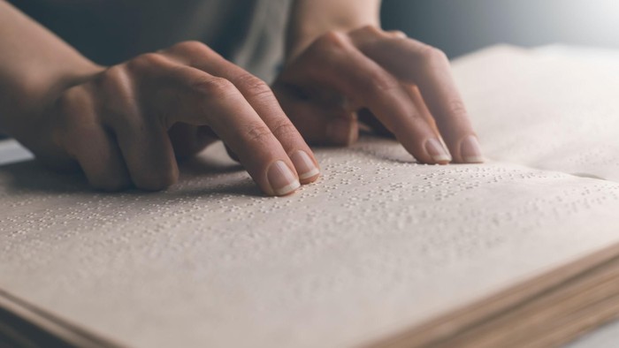 Eine Nahaufnahme zeigt Hände, die mit den Fingerspitzen aufmerksam über die erhabenen Punkte einer aufgeschlagenen Brailleschrift-Seite gleiten.