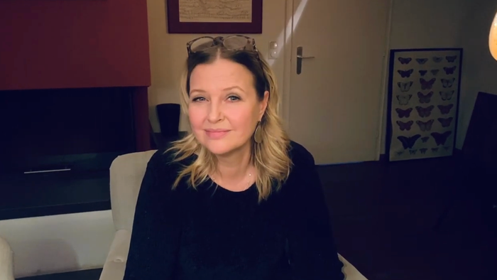 Eine Person mit blonden Haaren und Sonnenbrille sitzt drinnen in einem gemütlichen, schwach beleuchteten Raum.