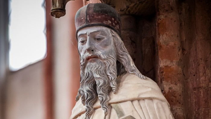 Statue einer bärtigen religiösen Figur, die ein Buch und einen Stab in einer historischen Umgebung hält.