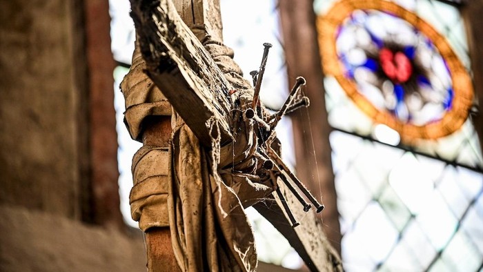 Ein verwittertes Holzkreuz mit Jesusfigur steht vor einem farbenprächtigen Kirchenfenster aus Buntglas.
