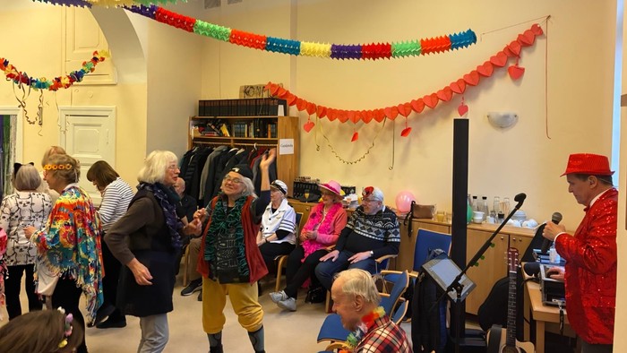 In einem bunt geschmückten Gemeindesaal tanzen und feiern verkleidete Seniorinnen und Senioren ausgelassen zu der Musik eines Alleinunterhalters, während andere fröhlich zuschauen und mitklatschen.