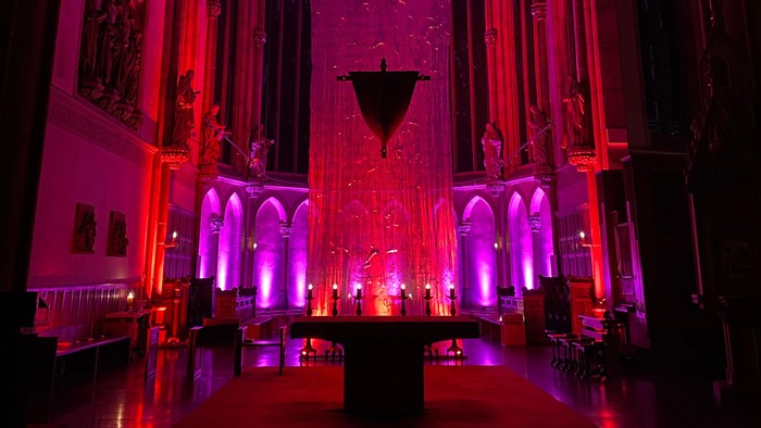Bild des Chorraumes in St. Pankratius in der Fastenzeit. Das große Kreuz, das von der Decke hängt, ist verhüllt und der Chorraum ist in rotes und violettes Licht getaucht.