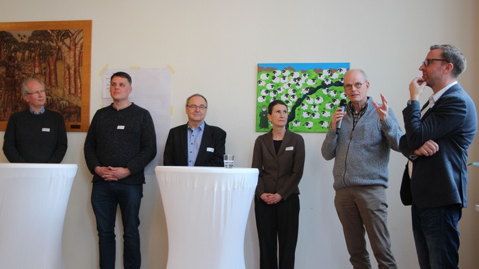 Fünf Personen stehen bei einer Podiumsdiskussion mit dekorativer Wandkunst und Namensschildern während einer Indoor-Veranstaltung.