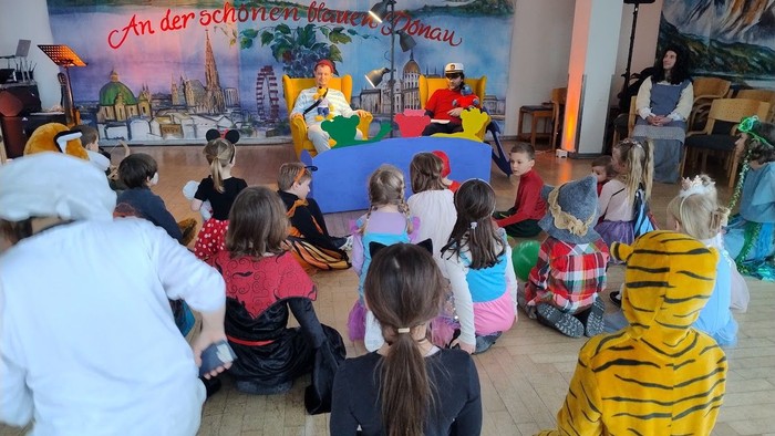Kinder in Kostümen schauen sich eine Puppenshow auf der Bühne mit farbenfroher Kulisse und einem Darsteller an.