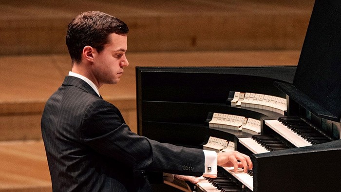 En mand i formelt tøj spiller på et orgel under en koncert i en træbygning.