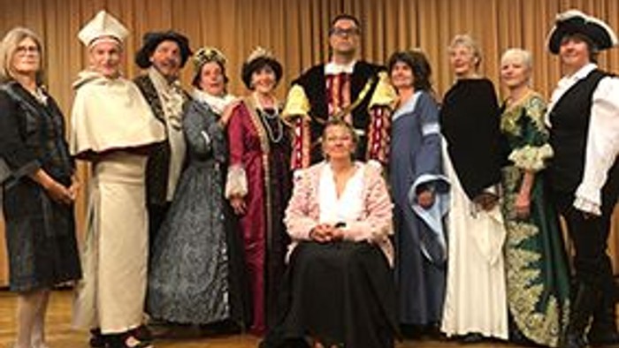 Etwa ein Dutzend Personen steht und sitzt auf einer Bühne vor einem geschlossenen Vorhang. Die meisten tragen aufwendige historische Kostüme, die an verschiedene Epochen erinnern: lange Gewänder, Mäntel, Hüte und festliche Kleider in unterschiedlichen Far
