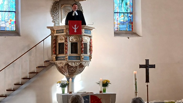 Ein Geistlicher predigt von einer verzierten Kanzel in einer Kirche, mit bunten Glasfenstern und sichtbaren Altarverzierungen.