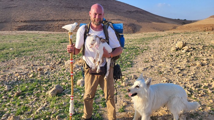 Ein Wanderer mit einer pinken Mütze trägt eine Katze, während er neben einem weißen Hund in einer wilden Outdoor-Landschaft steht.