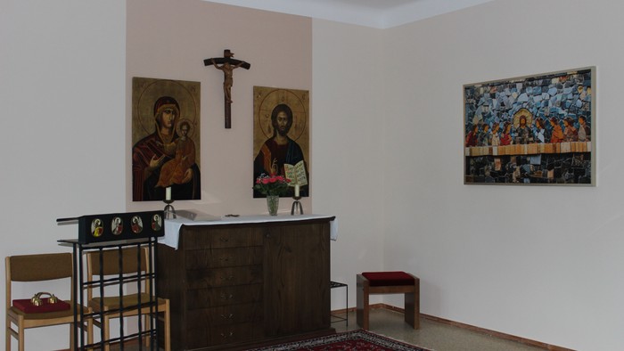 Eine ruhige Kapelle mit religiösen Ikonen, einem hölzernen Altar und schlichten Sitzgelegenheiten auf gemustertem Bodenbelag.