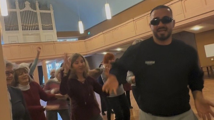 Seniorinnen und Senioren tanzen mit jungem Mann in Sonnenbrille zu modernen Beats im Großen Saal.