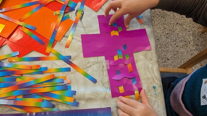 Ein Kind bastelt mit bunten Papierstreifen eine Kreuzform auf einem Tisch.