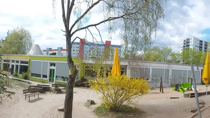 Freiluft-Spielplatz mit Bänken, Bäumen und modernen Gebäuden im Hintergrund.