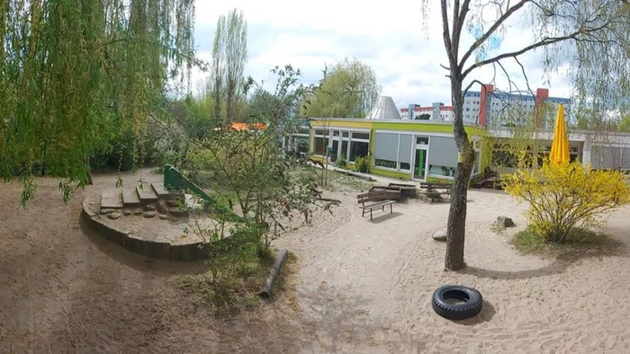 Freiluft-Spielplatz mit Bänken, Bäumen und modernen Gebäuden im Hintergrund.