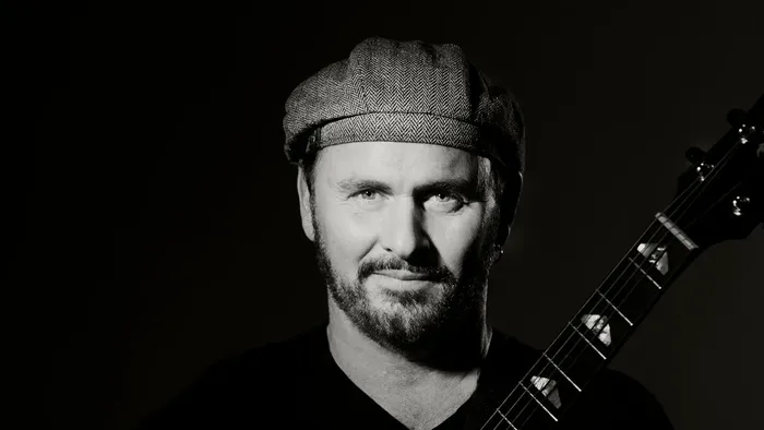 Ein Mann mit einer Mütze spielt eine akustische Gitarre vor dunklem Hintergrund.