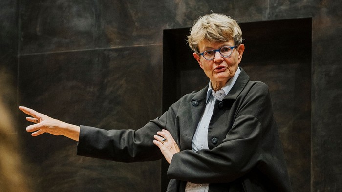 Ein Redner in einem Mantel gestikuliert, während er eine aufmerksame Zuhörerschaft drinnen anspricht.