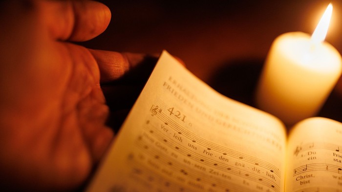 Symbolfoto: Im Kerzenlicht ist ein geöffnetes Gesangbuch zu sehen. Eine Hand hält es.
