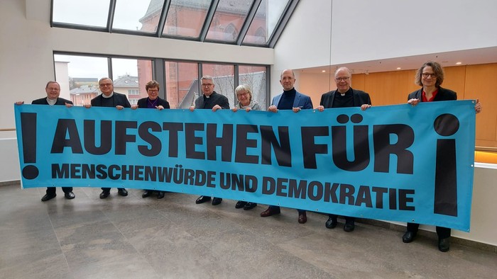 Acht erwachsene Personen stehen nebeneinander in einem hellen, modernen Innenraum mit großen Fensterflächen und Glasdach. Gemeinsam halten sie ein langes, türkisfarbenes Banner vor sich. Darauf steht in großen schwarzen Buchstaben: „AUFSTEHEN FÜR“ sowie d