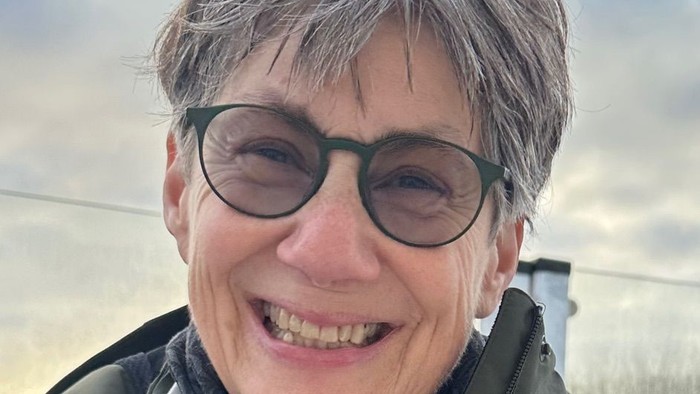 Eine fröhliche Person mit kurzem grauen Haar, die eine Brille und eine Jacke trägt und draußen lächelt.