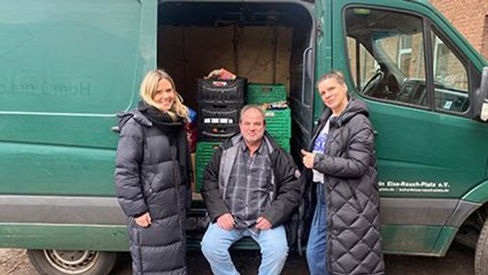 Drei Personen posieren in der Nähe eines offenen Lieferwagens, der mit Kisten voller Obst und Gemüse beladen ist.