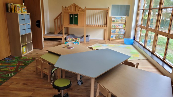 Das Bild zeigt einen Gruppenraum der neuen Kita mit Tischen, einem Schrank, einer Hochebene und einer großen Fensterfront. Kinder sind nicht zu sehen. 