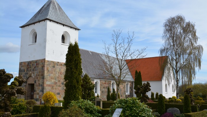 Hyggelig kirke med hvide mure, stensokkel og rødt tag omgivet af velplejet grønt og træer.