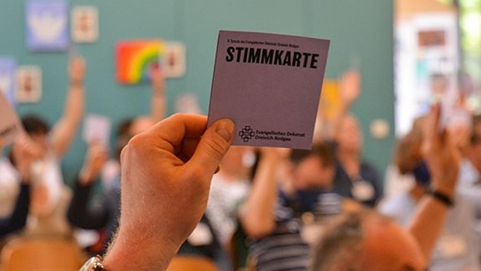 Eine erhobene Hand hält eine violette Stimmkarte mit der Aufschrift „Stimmkarte“ und dem Logo des Evangelischen Dekanats Dreieich-Rodgau. Im unscharfen Hintergrund sitzen zahlreiche Personen in einem Saal und heben ebenfalls ihre Stimmkarten zur Abstimmun