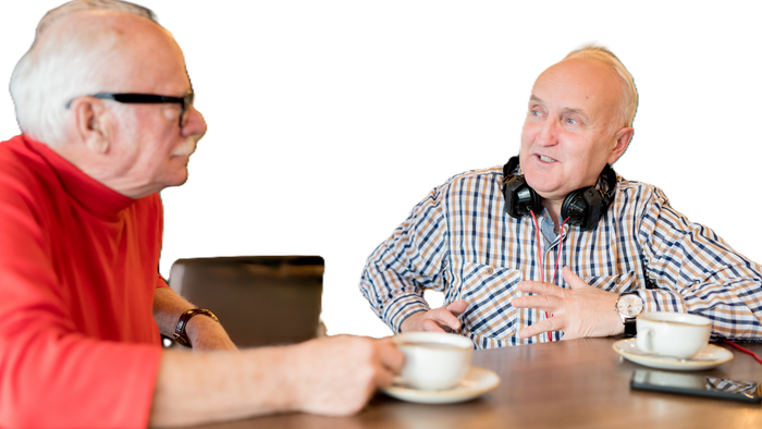 Zwei Männer in legerer Kleidung unterhalten sich bei einer Tasse Kaffee an einem Tisch in einer schwach beleuchteten Umgebung.