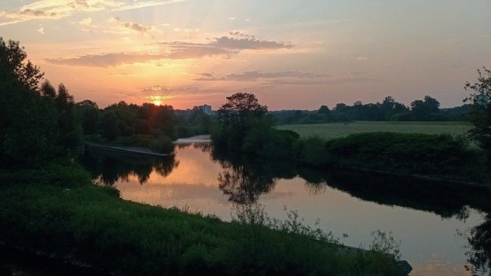 Ruhiger Sonnenuntergang, der sich über einen stillen Fluss spiegelt, umgeben von üppigem Grün und fernen Feldern.