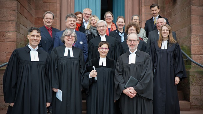 Eine größere Gruppe von Frauen und Männern steht auf den Stufen vor einem Kirchenportal aus rötlichem Sandstein. Im Hintergrund sind eine hohe Tür in kräftigem Blau sowie Teile der Kirchenfassade zu sehen. Im Vordergrund stehen mehrere Personen in schwar
