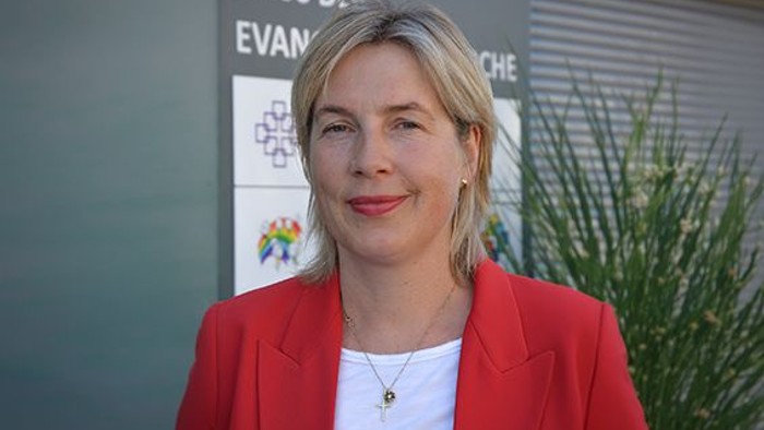 Professionelle Frau in rotem Blazer, die vor dem Schild „Haus der Evangelischen“ im Hintergrund lächelt.