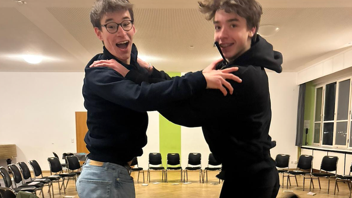 Zwei Menschen umarmen sich freudig und heben einander in einem runden Raum voller Stühle hoch.