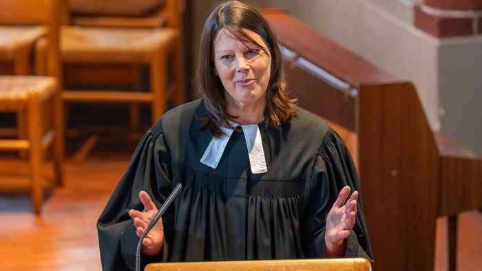 Ein Richter in schwarzer Robe spricht an einem Podium in einem Gerichtssaal mit hölzernen Kirchenbänken und Säulen.