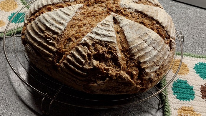 Frisch gebackenes Sauerteigbrot mit Kreuzschnitten auf einem Kühlgitter neben einer Backschüssel und einem Ofen.