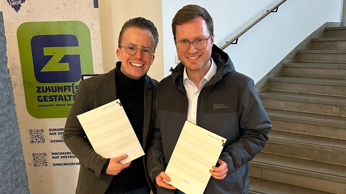 Zwei Männer halten Dokumente vor einer Treppe mit einem Kampagnenbanner „Zukunft gestalten“.