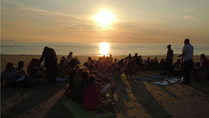 Gruppe versammelt sich am Strand bei Sonnenuntergang, sitzend und stehend um ein offenes Feuer am Meer.
