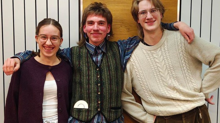 „Drei Personen stehen eng beieinander in einem Innenraum, jede Person legt einen Arm um die Schulter der danebenstehenden Person. Die Person auf der linken Seite trägt eine dunkle Strickjacke über einem hellen Oberteil. Die Person in der Mitte trägt ein g