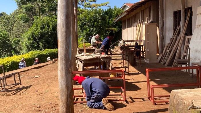 Mehrere Personen arbeiten im Freien neben einem Holzgebäude an Werkbänken und Holzkonstruktionen auf einem sonnigen, ländlichen Gelände.