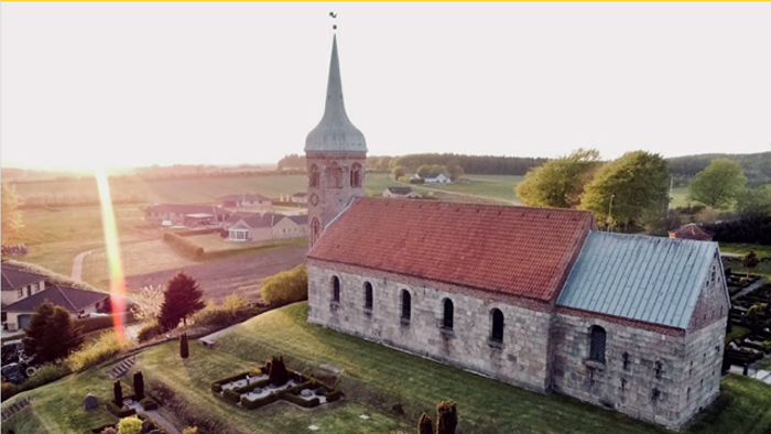 Luftfoto af en idyllisk stenkirke med rødt tegltag og en fredfyldt kirkegård i et landligt landskab.