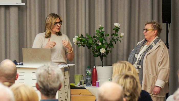 Zwei Frauen sprechen bei einer öffentlichen Veranstaltung vor einem aufmerksamen Publikum und einem blumengeschmückten Mittelstück auf der Bühne.