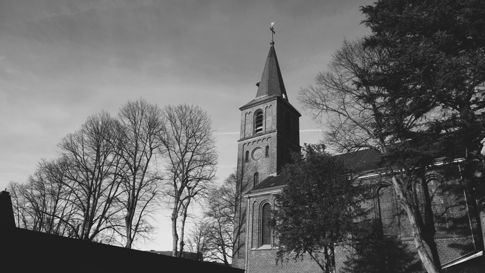 Ein markanter Schwarz-Weiß-Blick auf einen Kirchturm, gerahmt von kahlen Bäumen und einem bewölkten Himmel.