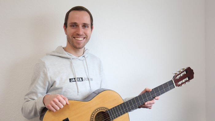 Ein lächelnder Mann hält und posiert mit einer hellen Akustikgitarre vor einer schlichten Wand.