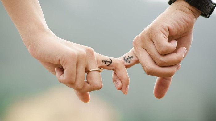 Zwei Hände, die Finger ineinander verschränkt halten, jede mit einer Tätowierung aus gekreuzten Ankern und einem Ring an einem Finger.