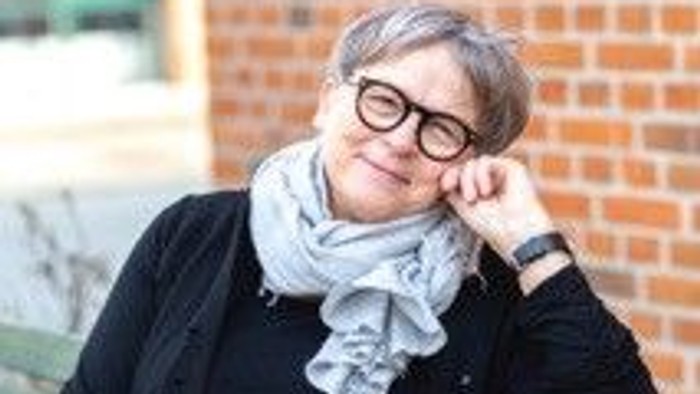 En kvinde med briller og tørklæde sidder eftertænksomt på en grøn bænk udendørs.