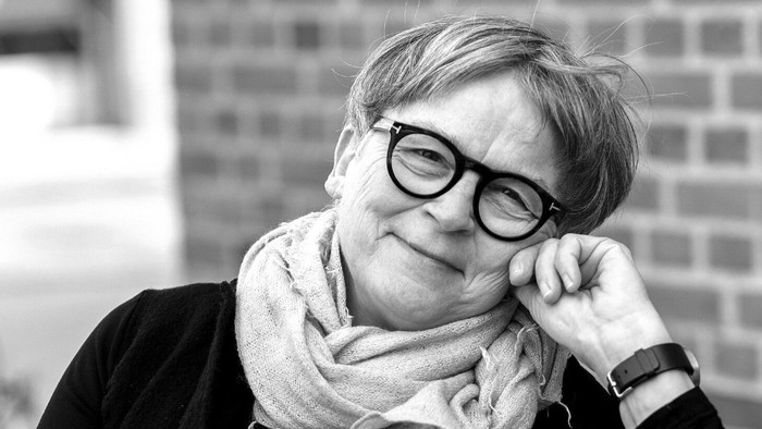 En glad kvinde med briller og et tørklæde, der støtter hovedet på hånden udendørs.