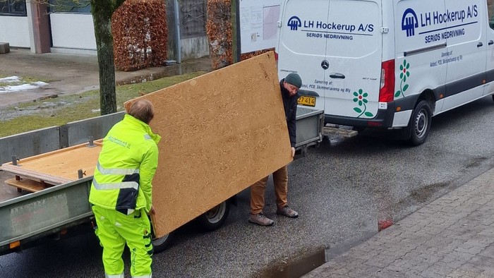 Arbejdere læsser store træpaneler på en leveringslastbil i et boligområde.