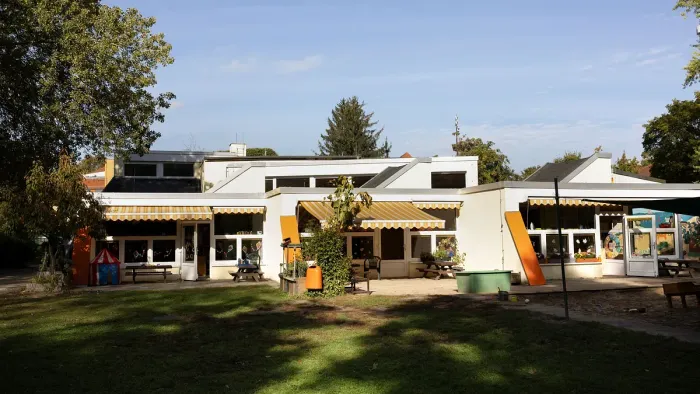 Modernes Gebäude mit orangefarbenen Akzenten, Außenbestuhlung und einer grasbewachsenen Fläche, umgeben von Bäumen.