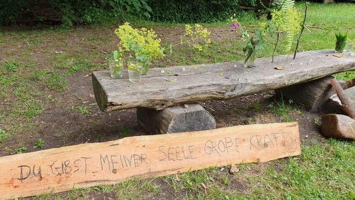 Eine Holzbank mit selbstgepflückten Blumen darauf. Ein Holzschild steht davor mit der Schrift: Du gibst meiner Seele große Kraft.