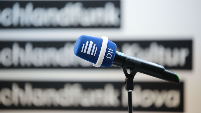 Ein blaues Mikrofon mit Deutschlandfunk-Logo steht auf einem Stativ vor unscharfem Hintergrund und symbolisiert eine Radiosendung oder Interviewaufnahme.
